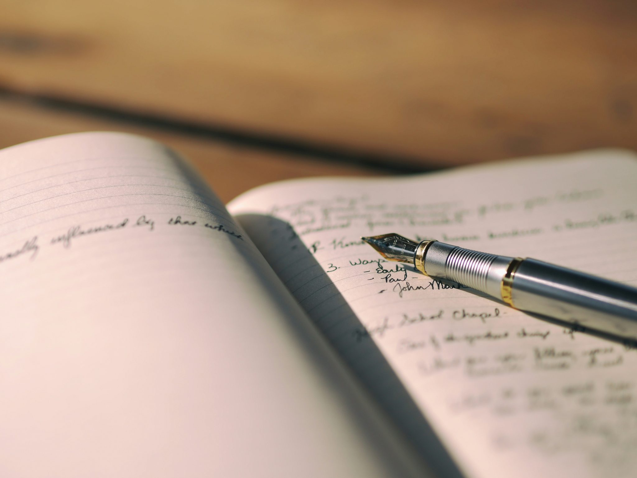 A close-up view of an open notebook with handwritten notes and a silver fountain pen resting on the pages. The background is softly blurred, focusing on the writing.